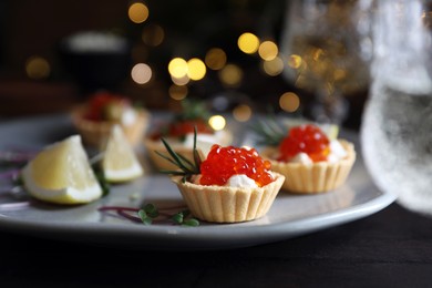Photo of Delicious tartlets with red caviar, cream cheese and lemon served near wine on black table, closeup. Space for text
