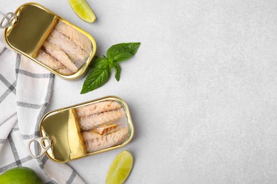 Photo of Open tin cans with mackerel fillets, lime and basil on grey table, flat lay. Space for text