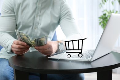 Online shopping. Cart illustration near laptop and man counting money indoors