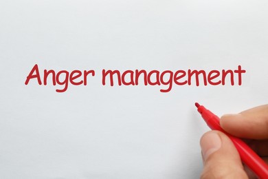 Photo of Man writing phrase Anger Management with red felt tip pen on white paper, top view