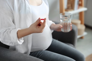 Pregnant woman holding pill and glass with water indoors, closeup