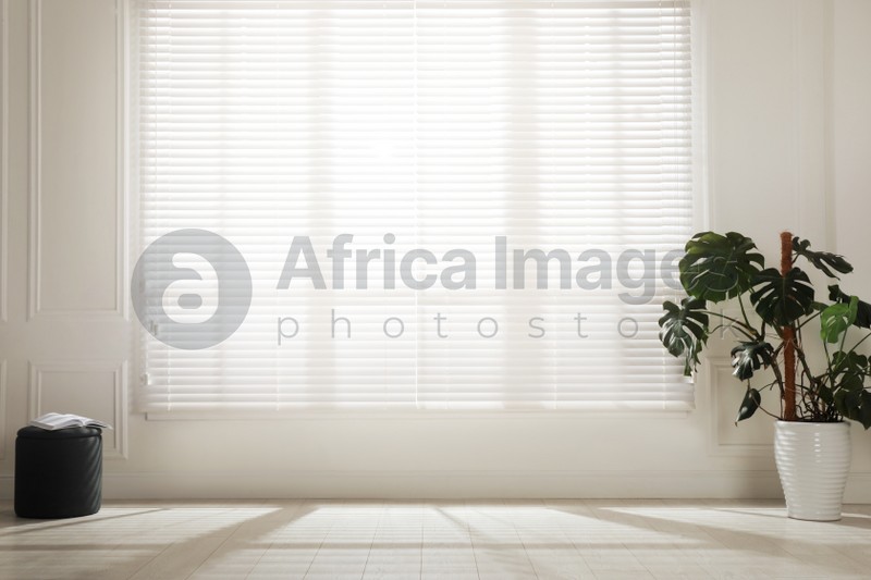 Photo of Monstera plant and pouf near large window with blinds in spacious room. Interior design