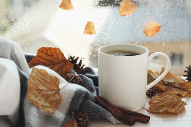 Cup of hot drink and autumn leaves near window on rainy day. Cozy atmosphere