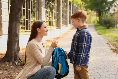 Photo of Young mom giving school backpack to her son outdoors