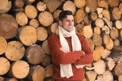 Handsome man wearing warm sweater and scarf near stack of firewood. Winter season