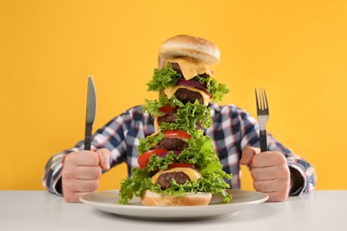 Hungry man with cutlery and huge burger at white table on yellow background