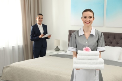 Chambermaid with stack of fresh towels and blurred supervisor in hotel room. Space for text