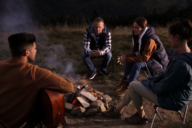 Group of friends gathering around bonfire at camping site in evening