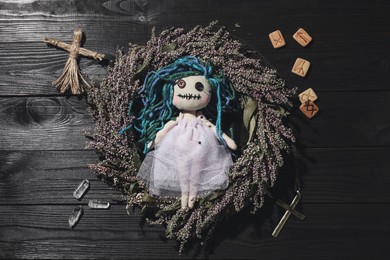 Female voodoo doll with pins surrounded by ceremonial items on black wooden background, flat lay