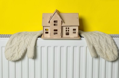 Wooden house model and knitted mittens on heating radiator near yellow wall. Energy efficiency concept