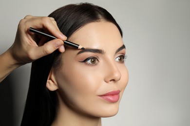 Artist correcting woman's eyebrow shape with pencil on grey background, closeup