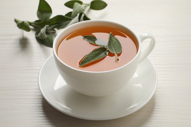 Cup of aromatic sage tea with fresh leaves on white wooden table