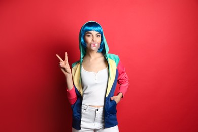 Fashionable young woman in colorful wig blowing bubblegum on red background