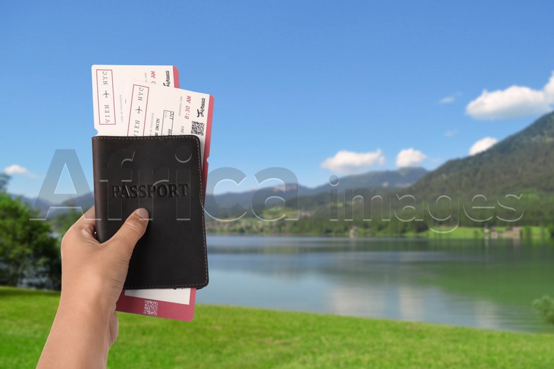 Woman holding passport with tickets near lake and mountains, closeup. Travel agency service