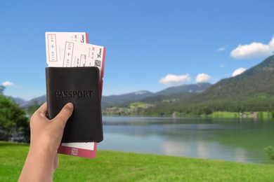 Woman holding passport with tickets near lake and mountains, closeup. Travel agency service