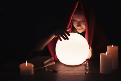 Photo of Soothsayer using glowing crystal ball to predict future at table in darkness, space for text. Fortune telling