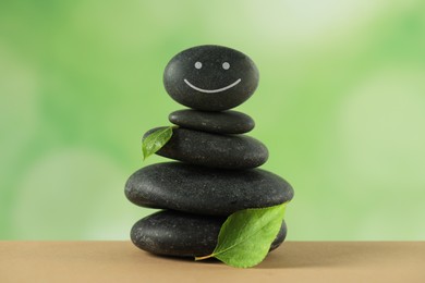 Photo of Stack of stones with drawn happy face and green leaves on table against blurred background. Zen concept