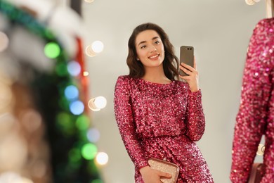 Photo of Beautiful young woman trying on stylish pink sequin dress and taking selfie near mirror in boutique. Party outfit