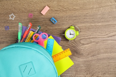 Stylish backpack with different school stationary and alarm clock on wooden table, flat lay. Space for text
