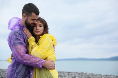 Young couple in raincoats enjoying time together under rain on beach, space for text