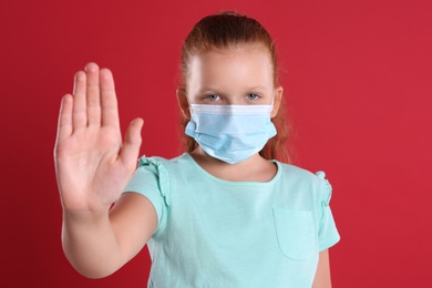 Little girl in protective mask showing stop gesture on red background. Prevent spreading of coronavirus