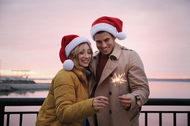 Photo of Couple in Santa hats holding burning sparklers near river