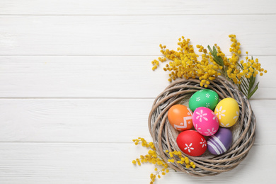 Colorful Easter eggs in decorative nest and mimosa flowers on white wooden background, flat lay. Space for text