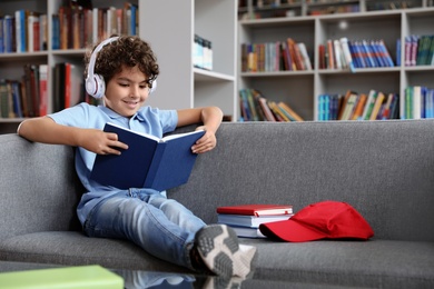Cute little boy in headphones reading book on sofa in library