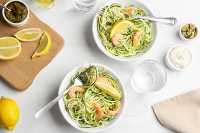 Delicious zucchini pasta with shrimps and lemon served on white wooden table, flat lay