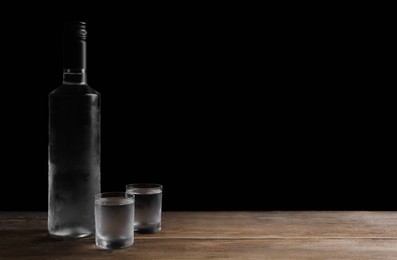 Photo of Bottle of vodka and shot glasses on wooden table against black background. Space for text