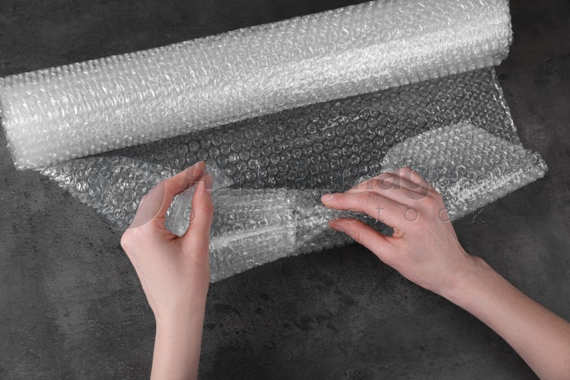 Photo of Woman covering wineglass with bubble wrap at dark grey table, closeup