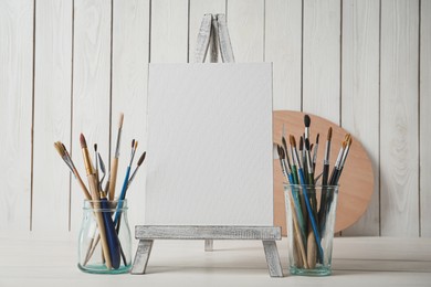 Easel with blank canvas and brushes on white wooden table