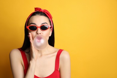 Fashionable young woman in pin up outfit blowing bubblegum on yellow background, space for text
