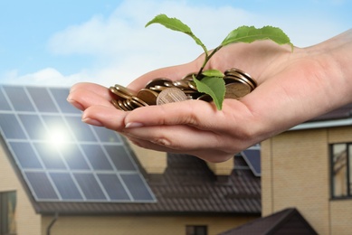 Woman holding coins and green sprout against house with installed solar panels on roof, closeup. Economic benefits of renewable energy