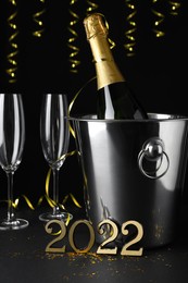 Happy New Year 2022! Bottle of sparkling wine in bucket and glasses on table against black background