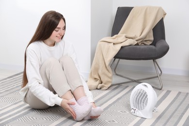 Photo of Young woman warming feet near electric fan heater at home