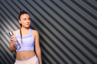 Young woman in sportswear with bottle near corrugated metal wall. Space for text