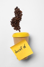 Note with word Decaf attached to takeaway cup and coffee beans on white background, flat lay