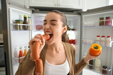 Young woman eating sausage near open refrigerator