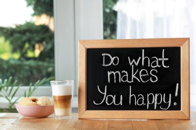 Blackboard with phrase Do What Makes You Happy, coffee and cookies on wooden table near window