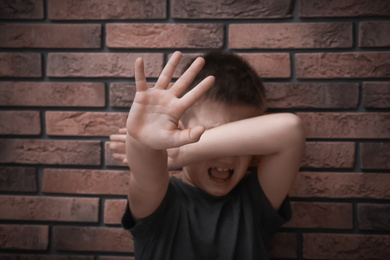 Photo of Scared little boy closing eyes with hand near brick wall. Child in danger