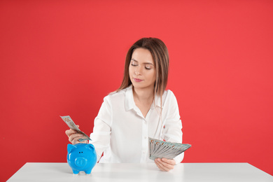 Young woman putting money into piggy bank at table on crimson background