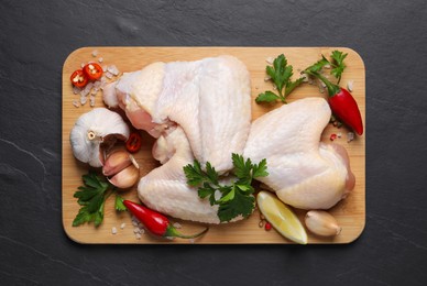 Wooden board with fresh raw chicken wings and other products on black table, top view