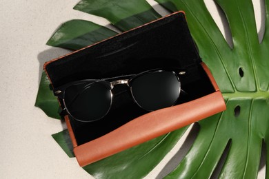 Stylish sunglasses in brown leather case and tropical leaf on sand, flat lay