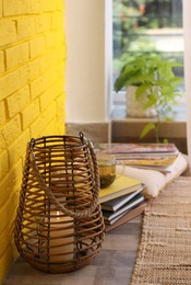 Stylish holder with burning candle and books on floor near yellow brick wall