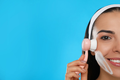 Young woman using facial cleansing brush on light blue background, space for text. Washing accessory