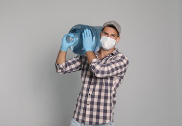 Courier in medical mask holding bottle for water cooler on light grey background. Delivery during coronavirus quarantine