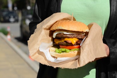 Woman holding delicious burger in paper wrap outdoors, closeup