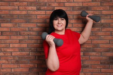 Happy overweight mature woman doing exercise with dumbbells near brick wall