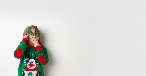 Cute little girl in Christmas sweater and party glasses on white background, space for text